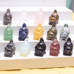 Puede incluir: Una colección de figuras de gato talladas en varios colores y materiales, incluyendo transparente, rosa, verde, azul, amarillo, marrón, negro y rojo. Los gatos se exhiben en una estantería blanca escalonada.