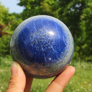 Op de afbeelding: Een gepolijste lapis lazuli bol, voornamelijk diepblauw met lichtere grijze en witte marmering. De edelsteenbal wordt in een hand gehouden, met een vage groene achtergrond van bladeren.
