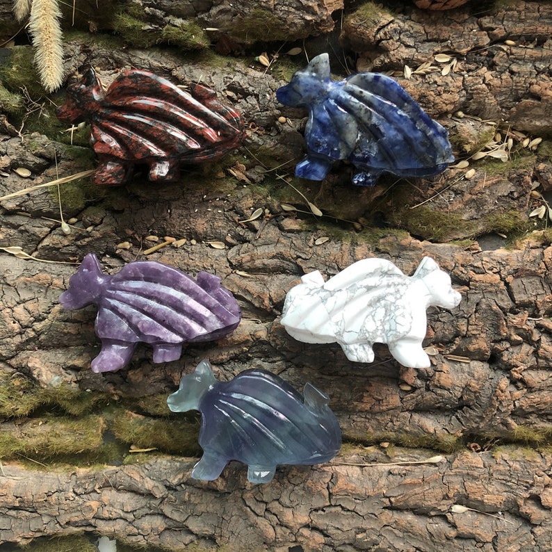 May include: A collection of carved stone dragon figurines in various colors. The dragons are red, blue, purple, white, and blue-green. The figurines are displayed on a textured, brown surface.