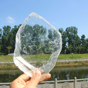 May include: A clear, irregularly shaped piece of selenite crystal is held up against a backdrop of trees, a lake, and a blue sky. The translucent crystal allows the background to be seen through it, creating a unique visual effect.