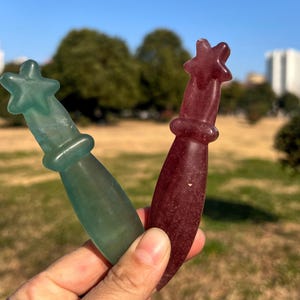 May include: Two gemstone wands, one teal and one burgundy, each with a star-shaped top and a rounded handle. The wands are held up against a blurred outdoor background, showcasing their polished, translucent appearance.