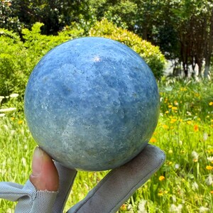 May include: A polished, spherical blue calcite crystal with a smooth, rounded surface. The stone displays a consistent light blue hue with subtle, natural mineral inclusions and a semi-translucent texture throughout its solid, circular form.