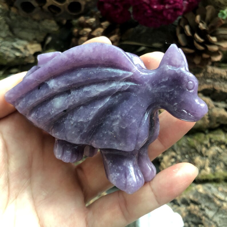 May include: A carved, purple gemstone dragon figurine. The dragon has detailed wings, a long neck, and four legs. The figurine is held in a hand, showcasing the intricate details of the dragon's form. The dragon is a fantasy-themed item.