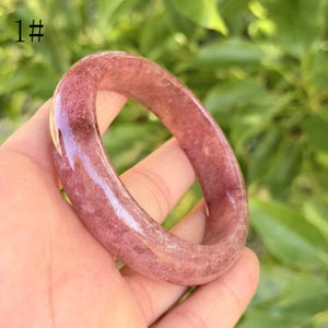 May include: A translucent, reddish-pink gemstone bangle bracelet. The smooth, polished surface reveals natural inclusions. The number "1#" is visible in the upper left corner. The bracelet is held in a hand against a blurred green background.