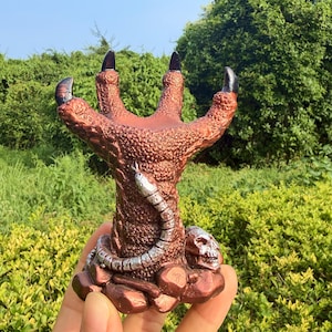 May include: A copper-colored sculpture shaped like a large, textured bird claw with four upward-pointing talons. A silver-colored snake wraps around the base, which is decorated with a small human skull and several rock-like formations.