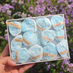 May include: A box of aragonite stones. Each stone is a light blue color and wrapped in clear plastic. The stones are labeled with small tan tags that read "ARAGONITE". The box is white and held in a person's hand.