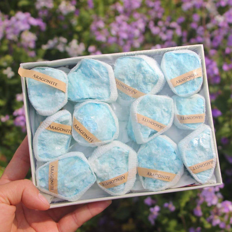 May include: A box of aragonite stones. Each stone is a light blue color and wrapped in clear plastic. The stones are labeled with small tan tags that read "ARAGONITE". The box is white and held in a person's hand.