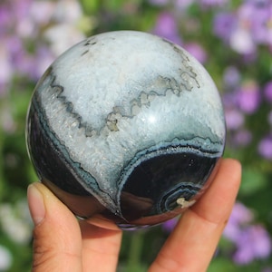 Puede incluir: Una esfera de ágata pulida con un llamativo patrón de bandas blancas, negras y verde azulado. La bola de piedra preciosa se sostiene en una mano, mostrando su belleza natural y sus intrincados detalles. La superficie de la esfera refleja la luz.