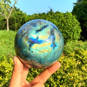 May include: A polished labradorite sphere featuring a smooth, rounded surface with iridescent flashes of blue, gold, and green. The stone displays natural internal fractures and deep colour variations that shift across the spherical form.