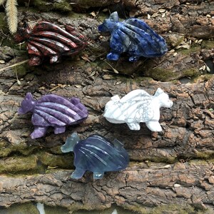 May include: A collection of carved stone dragon figurines in various colors, including red, blue, purple, white, and blue-green. The dragons are displayed on a textured, brown wooden surface, showcasing their detailed craftsmanship.