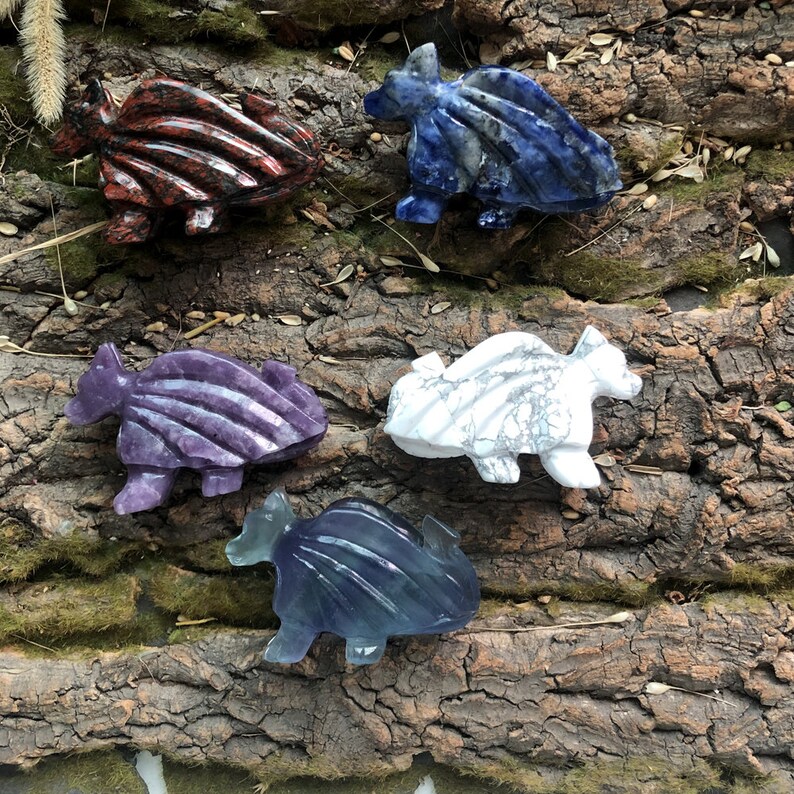 May include: A collection of carved stone dragon figurines in various colors, including red, blue, purple, white, and blue-green. The dragons are displayed on a textured, brown wooden surface, showcasing their detailed craftsmanship.