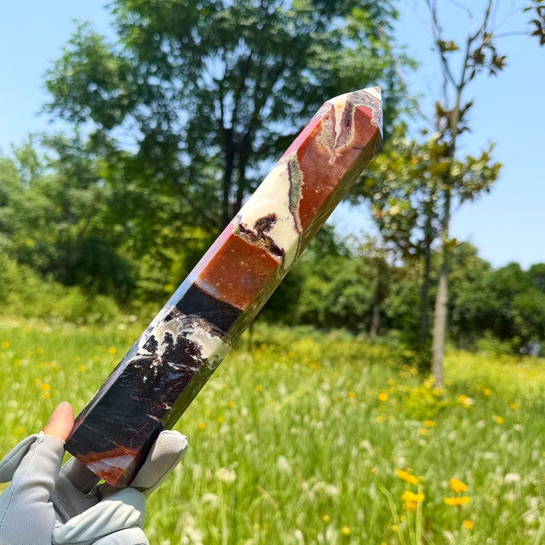 K&ouml;nnte beinhalten: Ein polierter, mehrfarbiger Steinturm mit einer hexagonalen Spitze. Der Kristall weist eine Mischung aus dunkelroten, dunkelbraunen, cremefarbenen und schwarzen Mineralmustern auf seinem l&auml;nglichen, facettierten K&ouml;rper auf und zeigt nat&uuml;rliche Farbvariationen.