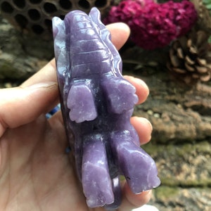 May include: A carved, lavender-colored elephant figurine. The sculpture is detailed with visible tusks, ears, and textured skin. The elephant is shown from a side angle, with its legs and trunk clearly visible. The figurine is held in a hand.
