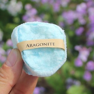 May include: A light blue aragonite crystal, held in a hand, with a tan paper band that reads "ARAGONITE". The crystal has a soft, fluffy texture. The background is blurred with green and purple foliage.