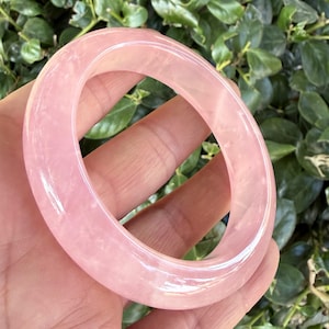 Könnte beinhalten: Ein glatter, durchscheinender Armreif aus Rosenquarz in hellem Rosa mit polierter Oberfläche und abgerundetem, kreisförmigem Profil. Das Edelsteinmaterial weist subtile interne Variationen und einen gleichmäßigen blassrosa Farbton auf.