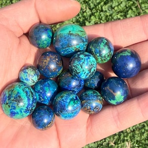 Peut inclure: Plusieurs sphères d'azurite polies, présentant un mélange de bleu profond et de turquoise vif. Les sphères, de tailles variées, sont tenues dans une main ouverte, avec un fond de feuillage vert.