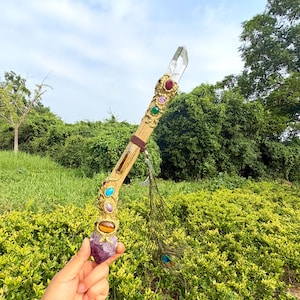 Può includere: Una bacchetta in legno con una punta di quarzo trasparente sulla sommità, decorata con filigrana in metallo dorato, gemme cabochon colorate incastonate, un avvolgimento in pelle marrone, un ciondolo a mezzaluna argentato e una lunga piuma di pavone.