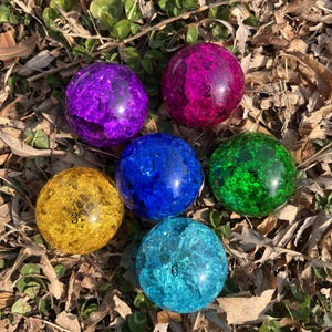 Puede incluir: Seis esferas de vidrio de colores, morado, rosa, amarillo, azul, verde y turquesa, están dispuestas sobre un lecho de hierba y hojas. Cada esfera tiene una apariencia agrietada y texturizada y está numerada del 1 al 6.