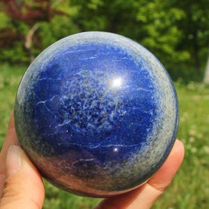 Op de afbeelding: Een gepolijste lapis lazuli bol. De diepblauwe steen heeft lichtere blauwe en grijze marmering, met gouden vlekken. De bol wordt in een hand gehouden, met een vage groene achtergrond van bladeren.