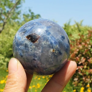Peut inclure: Une sphère de kyanite bleue avec des stries bleues et une inclusion brun foncé. La boule de cristal est tenue dans une main devant un fond de feuillage vert et de ciel bleu. C'est un objet décoratif.