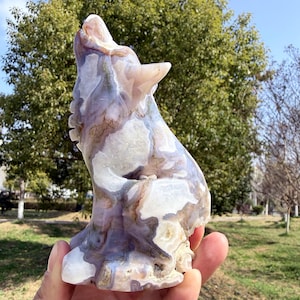 May include: A carved stone sculpture of a howling wolf. The wolf is depicted with its head raised and mouth open, showcasing a detailed design. The sculpture features a mix of white, purple, and brown colors, with a polished finish. The wolf statue is held in a hand.