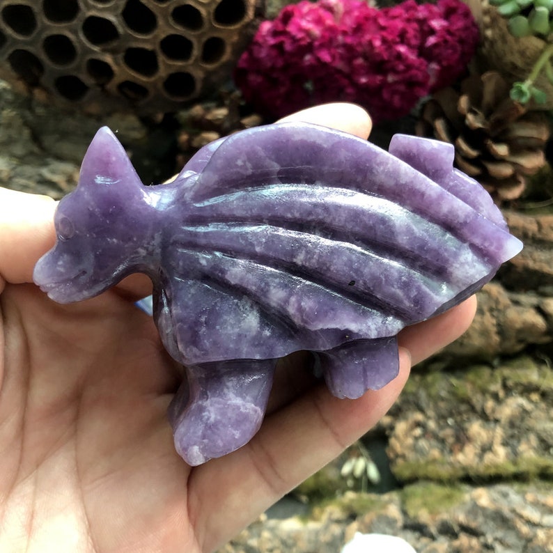 May include: A carved purple gemstone figurine of a dragon-like creature. The sculpture has a detailed design with wings and a spiky tail. The figurine is held in a hand, showcasing its size and intricate details. The dragon is a vibrant shade of purple.