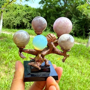 Puede incluir: Un soporte decorativo con una estructura en forma de árbol de color bronce con cinco piedras esféricas en tonos rosa pálido, gris y azul claro translúcido, todo montado sobre una base cuadrada negra para una presentación estable.
