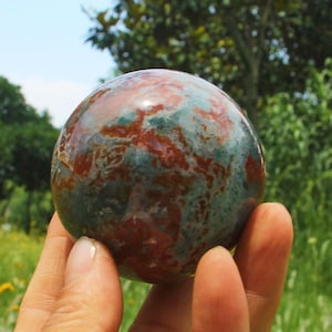 May include: A polished, spherical stone featuring a marbled pattern of deep forest green and earthy terracotta red. The surface is smooth and reflective, showcasing natural mineral inclusions that create a complex, organic, and multi-coloured texture.