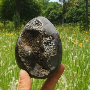 Puede incluir: Una piedra pulida en forma de huevo con una superficie exterior oscura y lisa. Un lado presenta una gran cavidad irregular llena de depósitos minerales oscuros y texturizados, con pequeñas formaciones cristalinas brillantes.