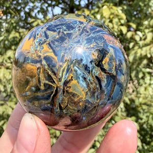 May include: A polished gemstone sphere with a swirling pattern of blue, gold, and black. The sphere is held in a hand, with a blurred green background of foliage. The gemstone has a reflective, glossy surface.