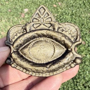 May include: A metallic gold-colored, eye-shaped decorative emblem with intricate detailing. The emblem features a central eye design, surrounded by ornate, crown-like embellishments. The piece has a textured surface and a dark, contrasting outline.