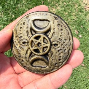May include: A gold-colored, circular pendant with intricate embossed designs. The pendant features a central five-pointed star within a circle, flanked by crescent shapes and stylized faces. The overall aesthetic suggests a mystical or symbolic theme.