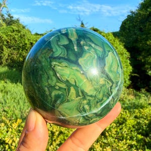 May include: A polished spherical stone featuring a marbled pattern of deep forest green, lime green, and creamy white swirls. The surface is smooth and reflective, displaying organic, flowing colour transitions across the entire round object.