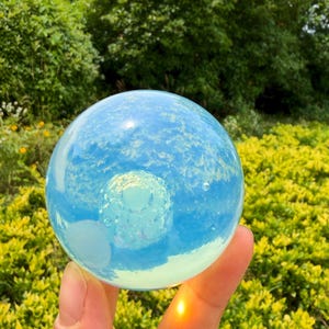 May include: A smooth, spherical glass orb with a translucent, pale blue hue. The surface of the sphere reflects the surrounding environment, creating a soft, iridescent glow. Inside the glass, a small, textured, circular pattern is visible.