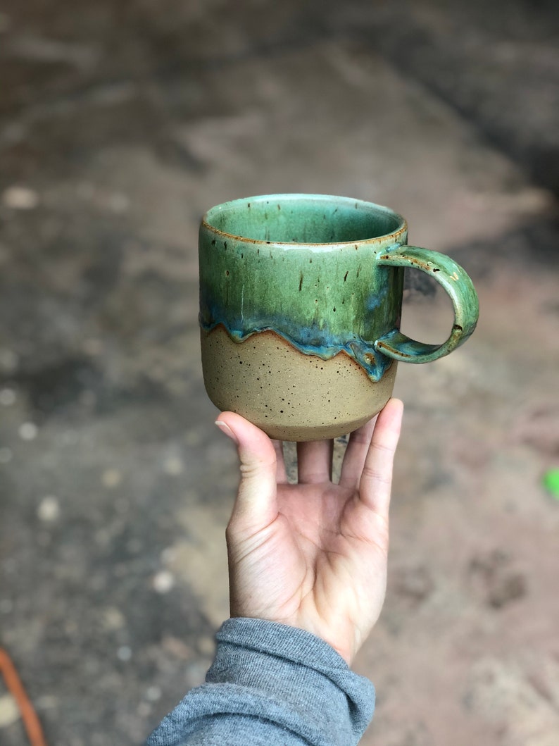 Peut inclure: Une tasse en c&eacute;ramique pr&eacute;sentant un design bicolore avec une base beige tachet&eacute;e et une gla&ccedil;ure verte brillante sur la partie sup&eacute;rieure. La gla&ccedil;ure coule sur les c&ocirc;t&eacute;s en un motif ondul&eacute;, et la tasse comprend une anse incurv&eacute;e.