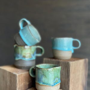 May include: Four handmade ceramic mugs with a blue and green glaze. The mugs are stacked on top of each other on a wooden surface.