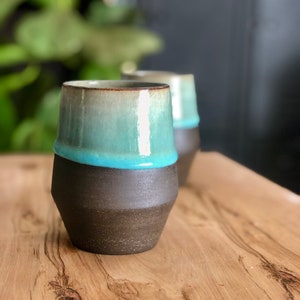May include: Two ceramic mugs with a brown glaze and a teal blue rim. The mugs are sitting on a wooden table.