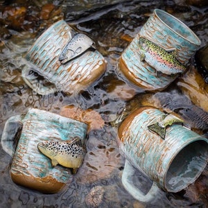 May include: Four ceramic mugs with fish designs. Each mug has a different fish species, including a trout, a salmon, and a brown trout. The mugs are sitting in a stream of water.