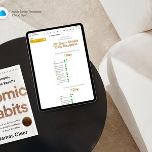 May include: A tablet displaying a 30-Day Daily Discipline chart sits on a black table next to a copy of "Atomic Habits" by James Clear. The book cover features gold lettering. The tablet's screen shows a habit tracker. The image is taken from above.