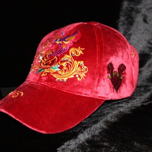 Puede incluir: Una gorra de béisbol de terciopelo rojo con diseños bordados de flores y pájaros. La gorra tiene una visera curvada y costuras negras contrastantes.