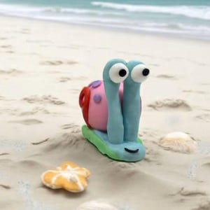 以下が含まれることがあります： ピンク色の体、青い眼柄、緑色の基部を持つ、気まぐれな粘土のカタツムリの彫刻。砂浜にヒトデと貝殻と一緒に置かれています。カタツムリは大きなギョロ目と笑顔の顔をしています。