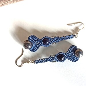 Peut inclure: Une paire de longues boucles d'oreilles en macramé bleu avec des crochets argentés et des perles gris foncé et violettes.