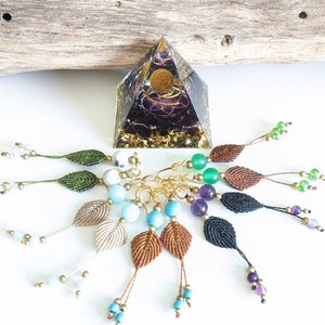 Peut inclure: Un ensemble de huit breloques en macramé en forme de feuille faites à la main avec des perles de différentes couleurs et un anneau en métal doré. Les breloques sont suspendues à une surface en bois.