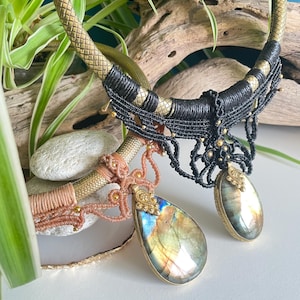 May include: Two gold and black macrame necklaces with iridescent gemstone pendants. The necklaces are layered on a wooden surface with green plants in the background.