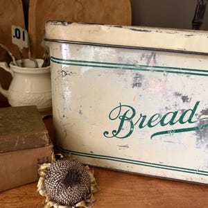 May include: A rectangular, cream-colored metal bread box with a removable lid, featuring green horizontal stripes and the word Bread in a green cursive script. The surface shows signs of wear, including paint chips and rust-like discoloration.