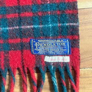 May include: A red, green, and black plaid wool scarf with fringe ends. A blue rectangular label reads: Warranted to be a Pendleton, Pendleton Woolen Mills, Pendleton, Oregon, 100% Virgin Wool. A white tag below reads: Sana Franke.