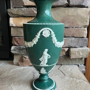 May include: A green ceramic vase with white relief designs. The vase has a round body with a narrow neck and a flared top. The design features a floral motif and a woman holding a flower.