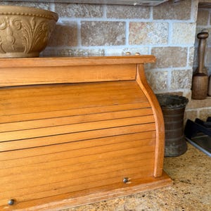 May include: A light brown wooden bread box with a roll-top tambour door and two small metal knobs. The bread box features a curved side profile and a flat top surface, providing a storage solution for baked goods in a kitchen setting.