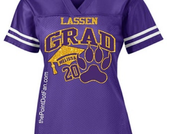 May include: Purple jersey with white stripes on the sleeves. The jersey has gold glitter text that says "Lassen Grad" with a graduation cap and paw print. The text "Melissa 20" is also on the jersey.