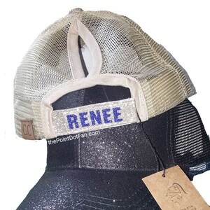 Pode incluir: Um bon&eacute; de beisebol preto com glitter, parte traseira em rede bege e abertura para rabo de cavalo alto. A tira traseira apresenta o nome RENEE em letras azuis brilhantes. Uma pequena etiqueta castanha est&aacute; presa ao lado, log&oacute;tipo C.C. vis&iacute;vel.